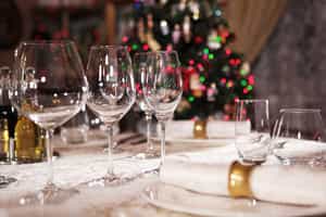 Copas de vidrio y platos sobre mantel blanco y árbol de Navidad con luces al prendidas al fondo